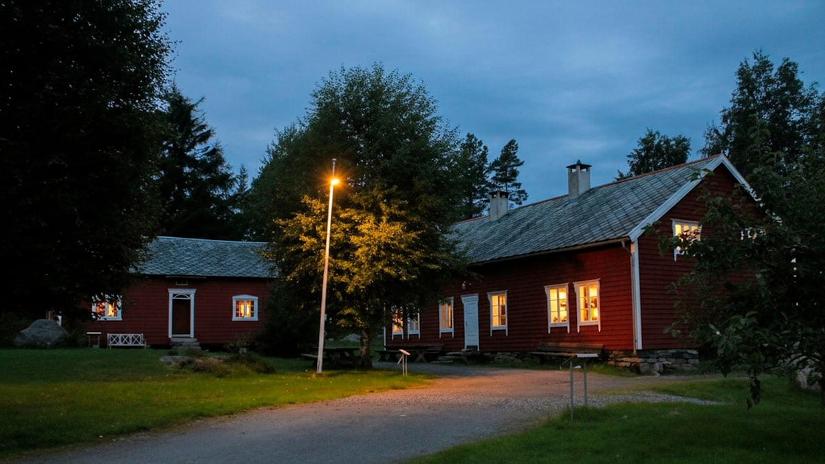 A charming evening scene featuring traditional red wooden farmhouses illuminated by warm interior lights in rural Norway. Surrounded by lush greenery and tall trees, the buildings stand beneath a moody blue-hour sky, while a solitary streetlamp adds a welcoming glow to the peaceful countryside setting. The photograph captures the cozy atmosphere and timeless character of Norwegian rural life.